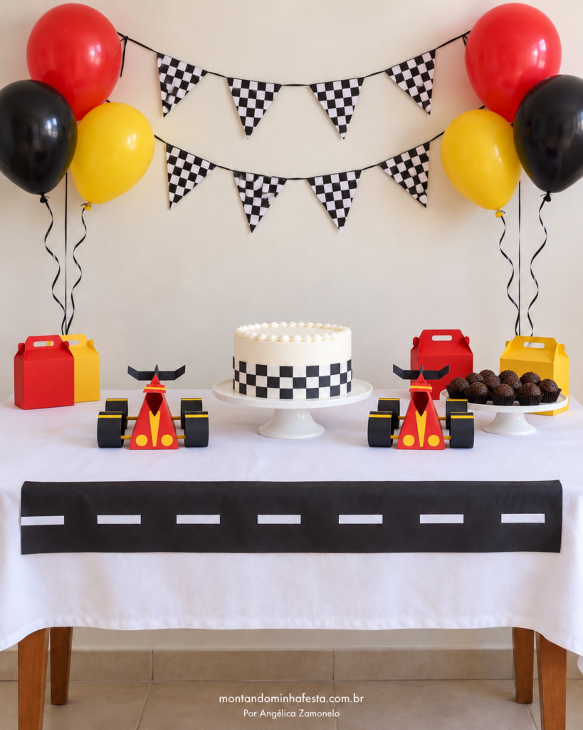 Mesa de festa vista de frente com dois carrinhos de papel vermelhos, pretos e amarelos, um de cada lado do bolo, do blog Montando Minha Festa.