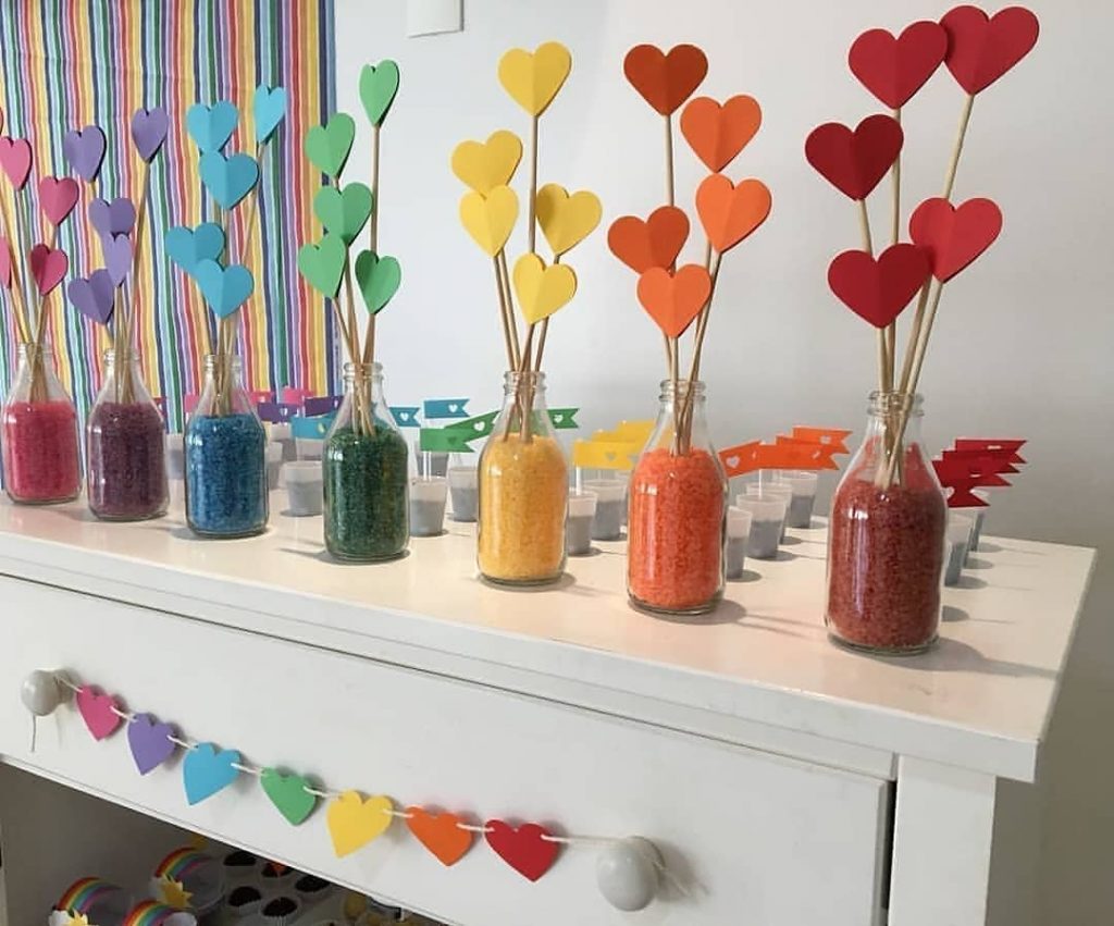 Centros de mesa com garrafas de vidro, sal colorido e corações de papel em cores do arco-íris, usados como decoração criativa para festas temáticas.