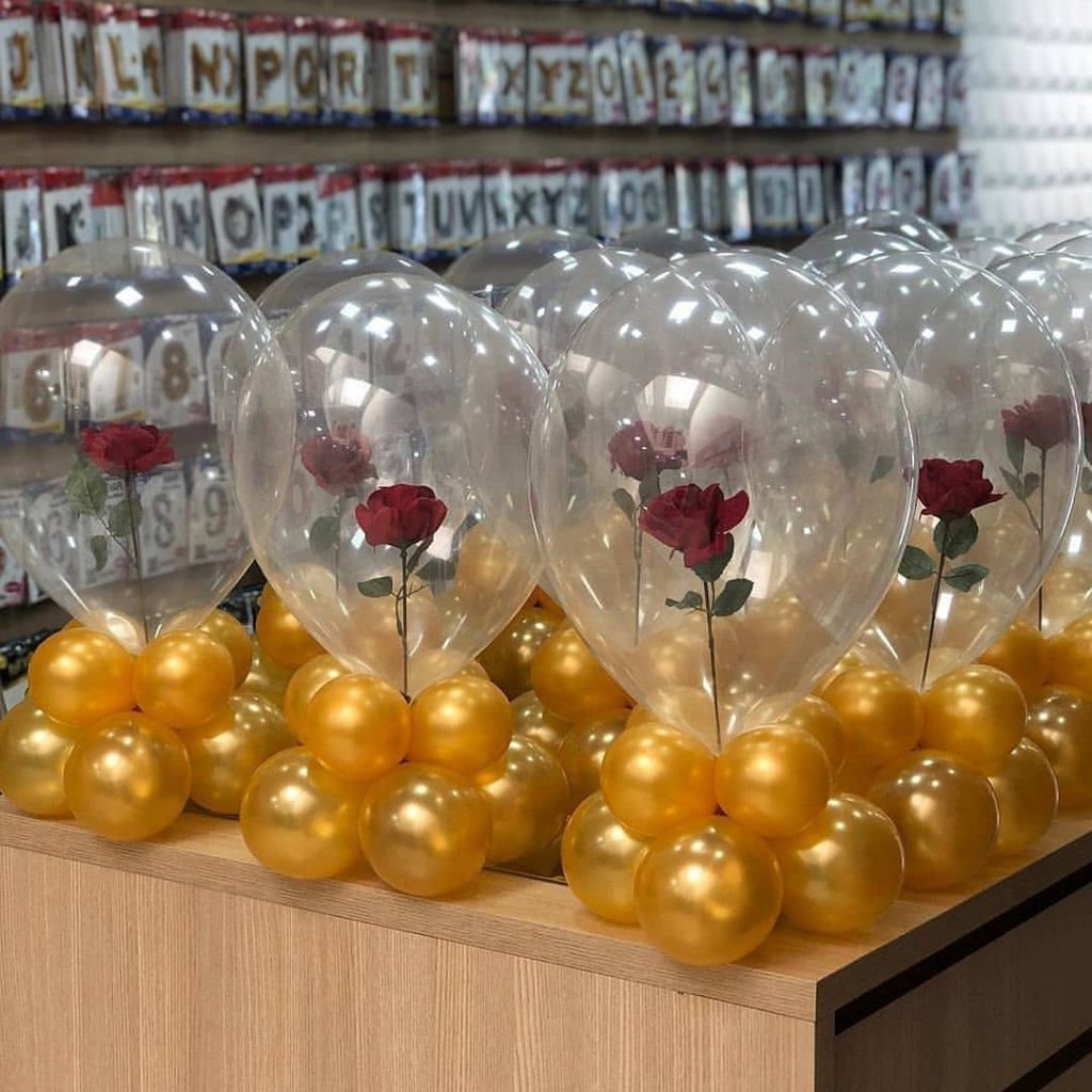 Centro de mesa com balão transparente contendo rosa vermelha, apoiado em base de balões dourados, ideal para festas temáticas e elegantes.
