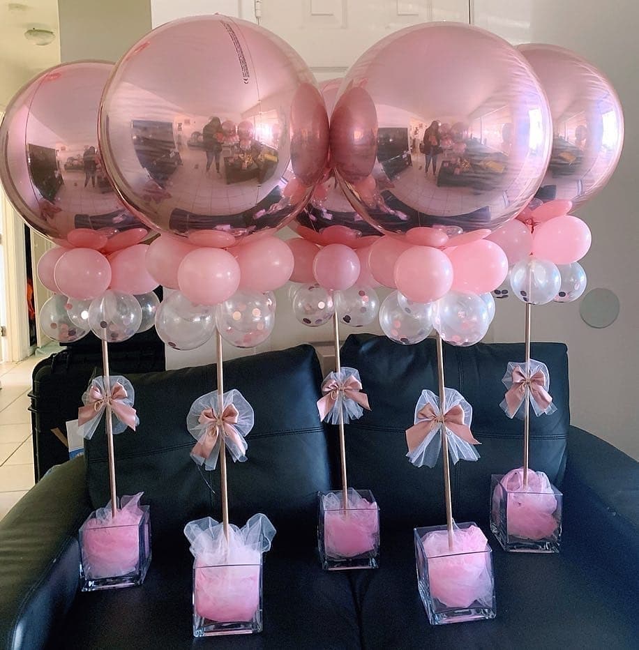 Centro de mesa com balões metalizados rosa e base de vidro decorada com tule e laço de cetim.