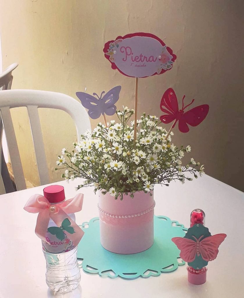 Centro de mesa com lata reutilizada pintada de rosa, decorada com flores do campo, topper com nome e borboletas em papel, usado em festa infantil personalizada.