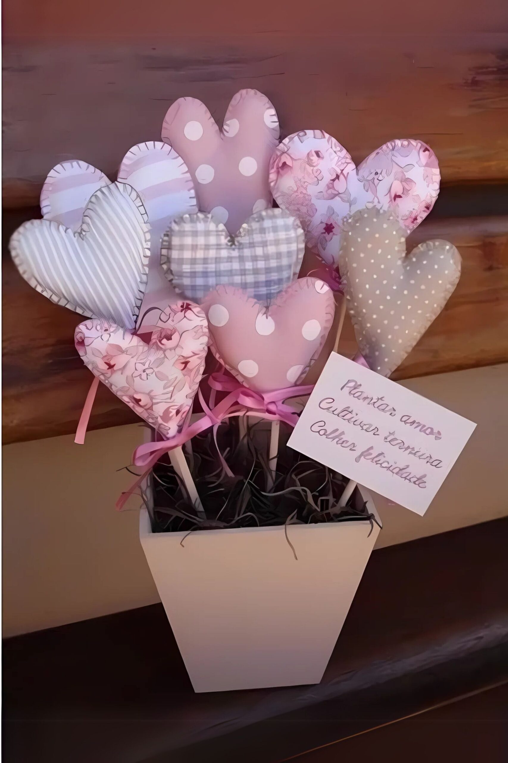 Centro de mesa com corações de tecido em tons rosa e bege, colocados em vasinho com musgo e plaquinha com mensagem de amor.