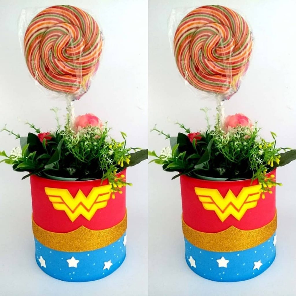 Centro de mesa com lata decorada em EVA tema Mulher Maravilha, com flores artificiais e pirulito colorido, ideal para festas infantis e super-heroínas.