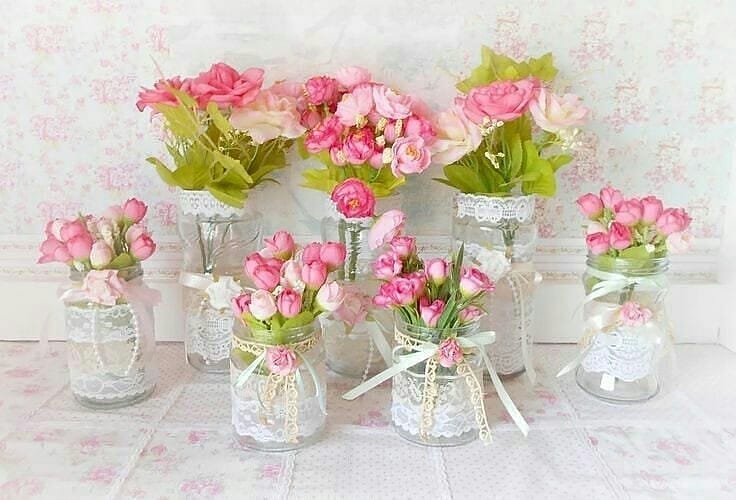 Centros de mesa com vidros decorados com renda, laços e flores rosa, ideais para casamentos românticos, festas vintage ou decoração estilo shabby chic.