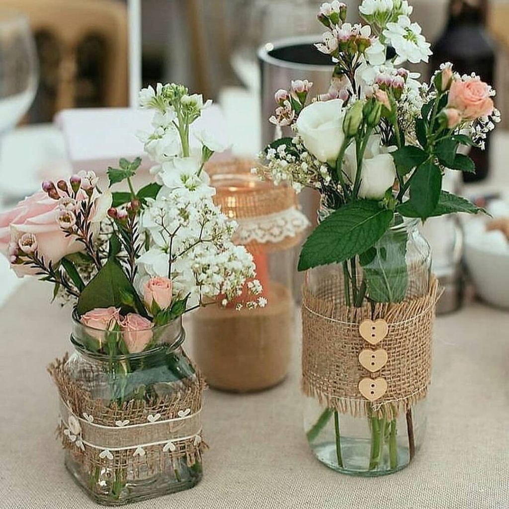 Centro de mesa rústico com vidros decorados com juta e botões, contendo arranjos de flores do campo em tons claros, ideal para casamentos e eventos ao ar livre.