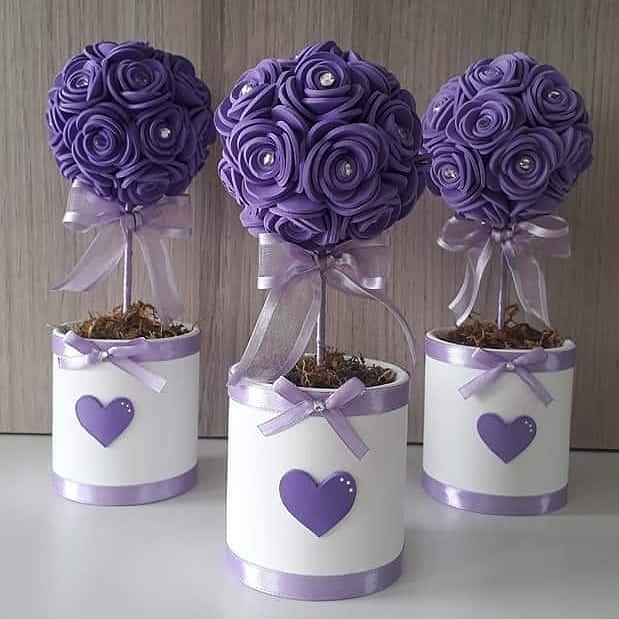 Centro de mesa com bola de rosas lilás em EVA, laço de fita e base branca decorada com coração em alto-relevo, ideal para festas elegantes e delicadas.