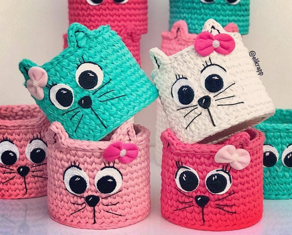 Cestinhas de crochê feitas com fio de malha em formato de gatinhos coloridos, com olhos grandes e laços decorativos, ideais para lembrancinhas ou decoração temática.