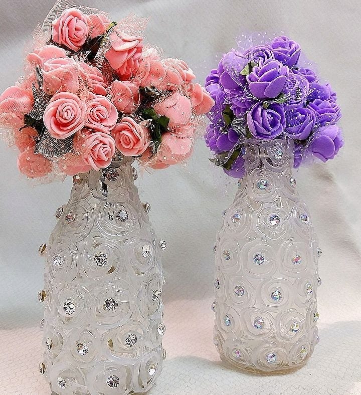 Garrafas artesanais decoradas com desenhos de cola quente e strass, usadas como centro de mesa com flores artificiais em rosa e roxo.
