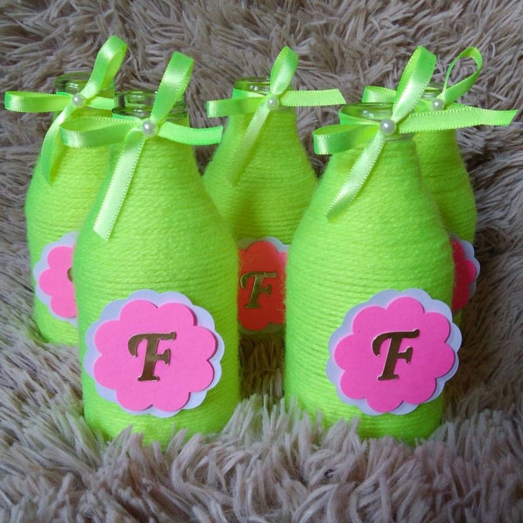 Garrafas decoradas com linha neon verde, laço de fita e aplique de flor com letra personalizada, usadas como centro de mesa ou lembrancinha de festa.