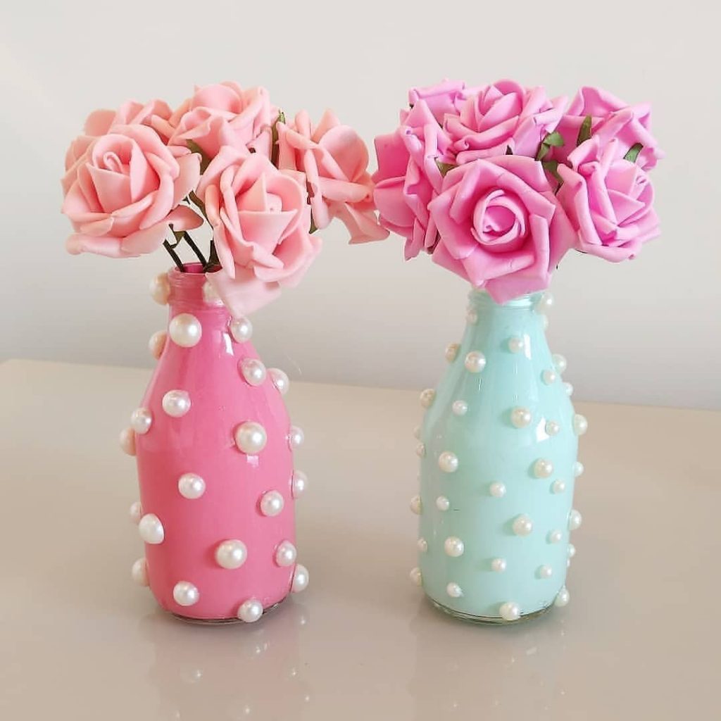Garrafas decoradas com tinta pastel e pérolas adesivas, usadas como centro de mesa com flores artificiais cor-de-rosa para festas delicadas.