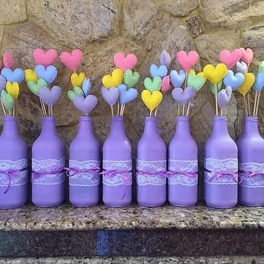 Garrafas lilás decoradas com renda e corações de feltro coloridos, usadas como centro de mesa para festas infantis e eventos delicados.