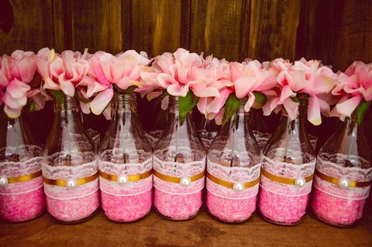 Garrafinhas decoradas com renda branca, fita dourada e flores rosas artificiais, usadas como centro de mesa delicado para festas femininas.