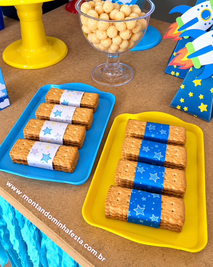 Biscoitos embalados com cintas de papel decoradas com estrelas azuis para festa Astronauta, organizados em bandejas azul e amarela, com lembrancinhas de foguete ao fundo.