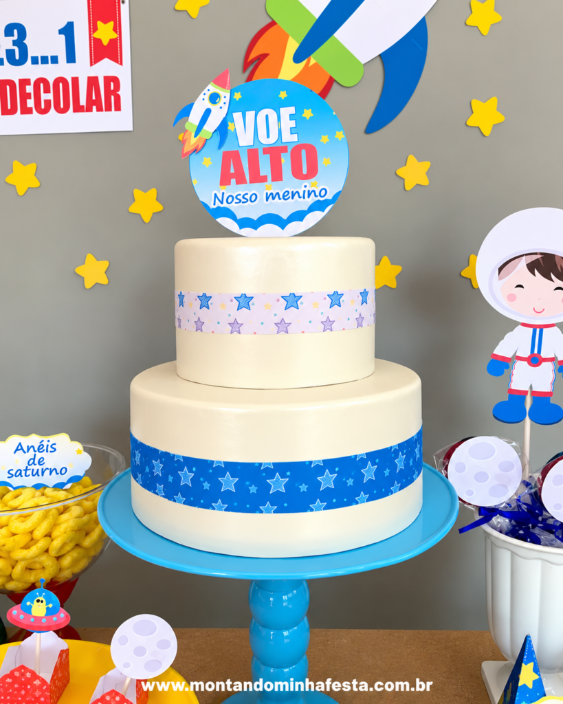 Bolo de dois andares para festa Astronauta, com cobertura branca, faixas azuis com estrelas, topo de bolo com foguete e frase Voe Alto, Nosso Menino, em decoração de aniversário infantil.