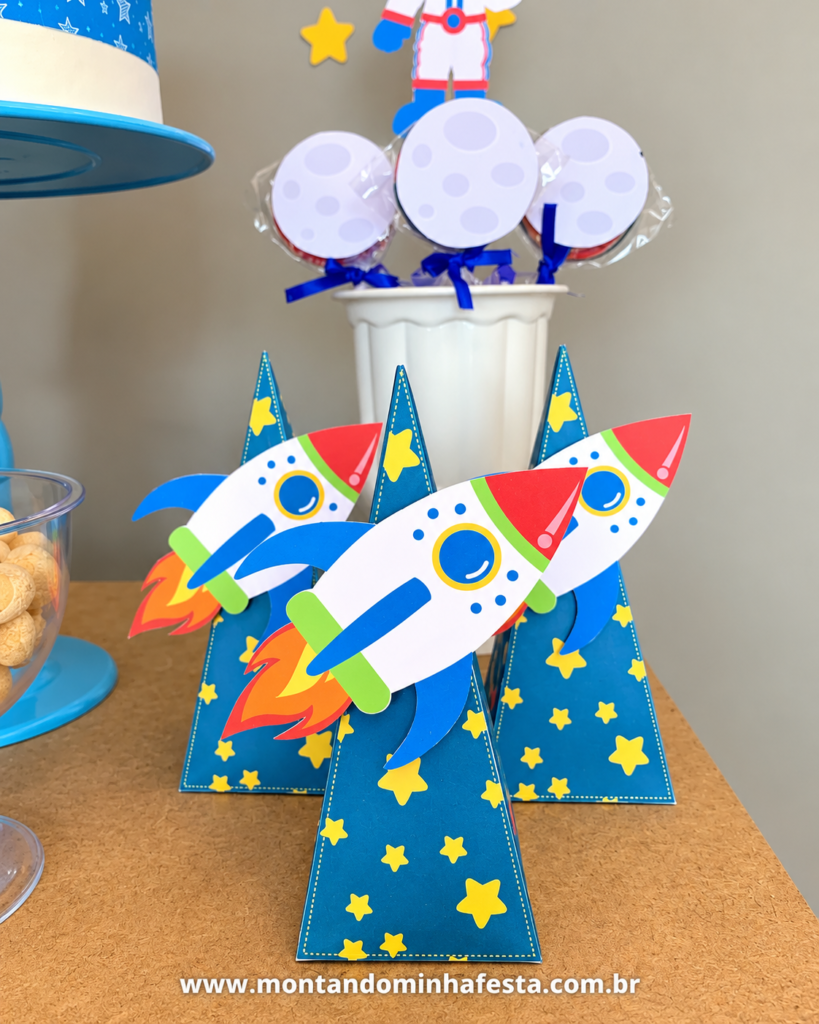 Três caixas pirâmide azuis com estrelas amarelas e apliques de foguetes coloridos, usadas como lembrancinhas de festa Astronauta, com pirulitos personalizados de lua ao fundo.