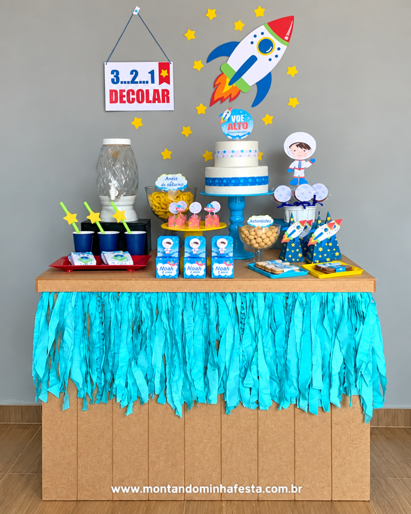 Decoração de festa Astronauta simples e barata, com mesa de madeira, saia de papel azul, bolo temático, foguete na parede, estrelas amarelas e lembrancinhas personalizadas para aniversário infantil.