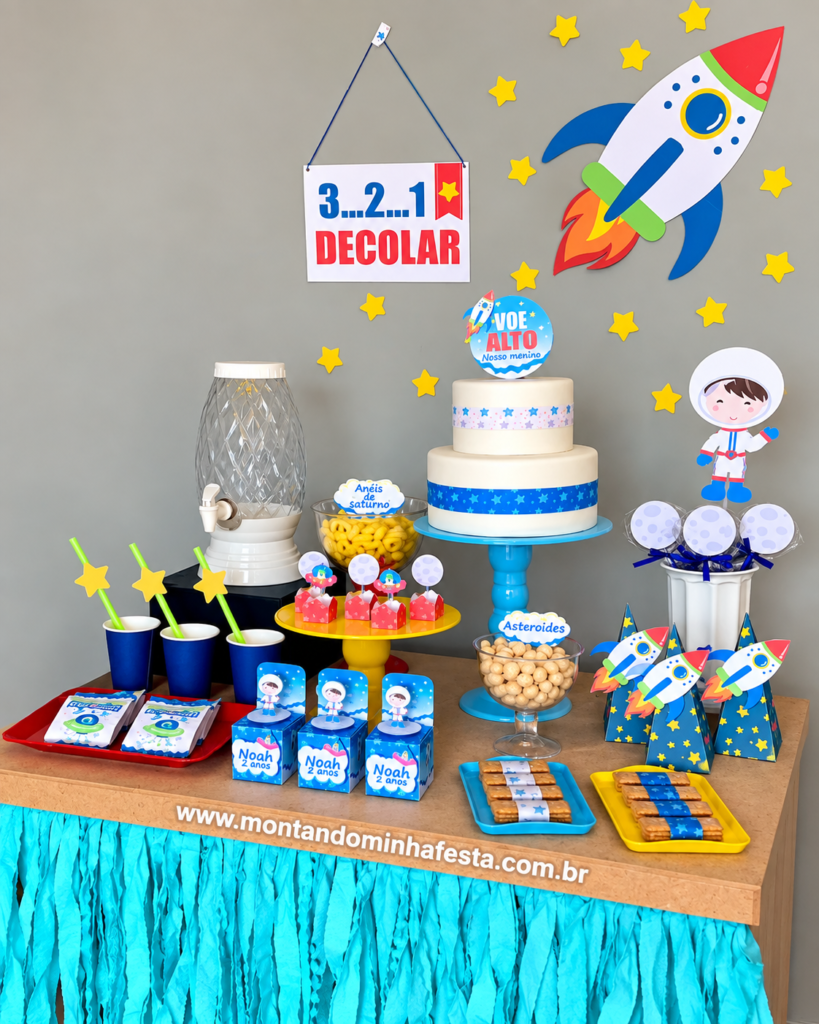 Mesa de festa Astronauta simples e barata, com bolo de dois andares, foguete e estrelas na parede, lembrancinhas personalizadas, docinhos temáticos e saia de papel azul.