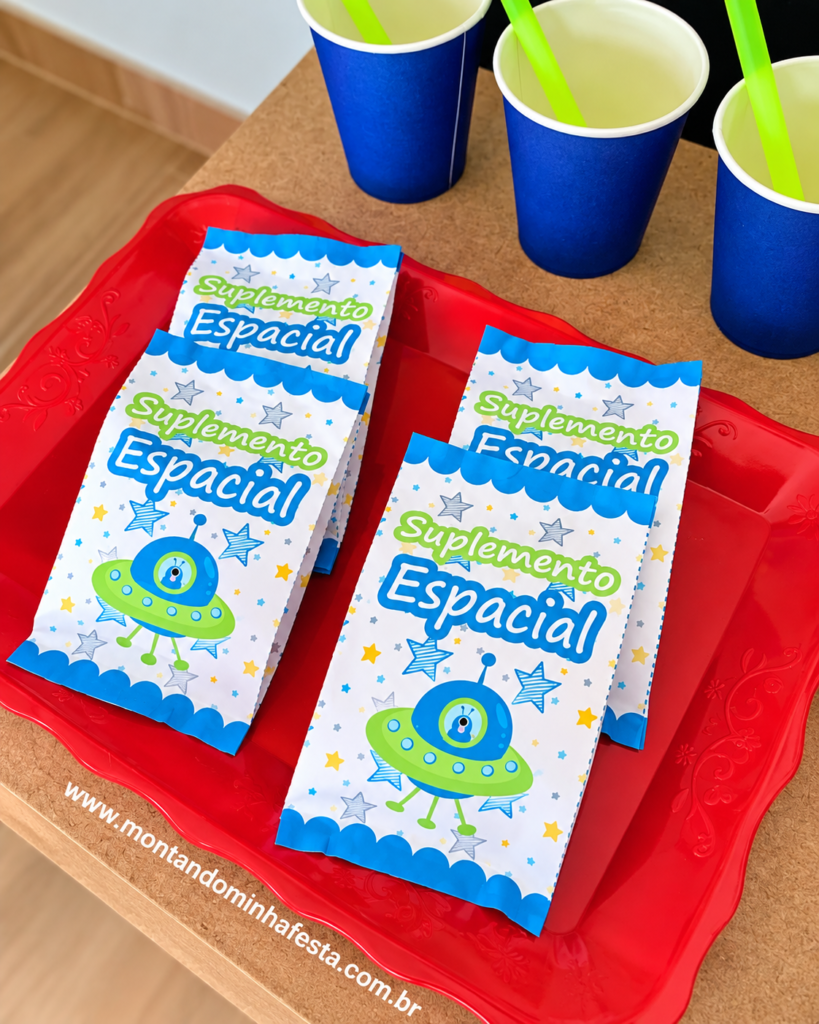 Embalagens personalizadas de Suplemento Espacial para festa Astronauta, decoradas com estrelas e disco voador, organizadas em bandeja vermelha com copos azuis ao fundo.